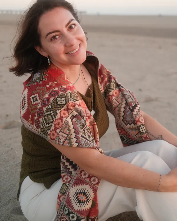 Nicole Sanchez, founder of Rooted Magic, smiling on the beach at golden hour in a patterned shawl