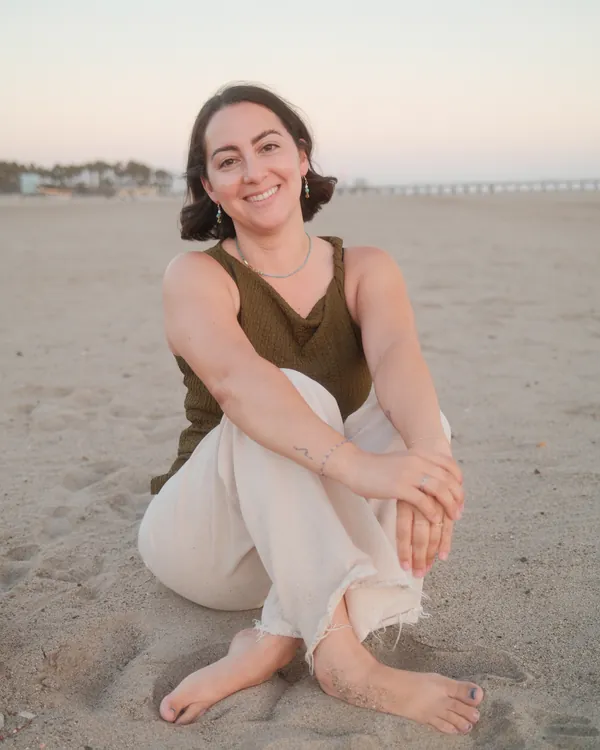Nicole Sanchez sitting barefoot in the sand, smiling, with the beach behind her